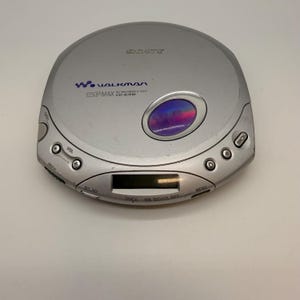 Può includere: Lettore CD Sony Walkman argentato. Il dispositivo ha un design circolare con un display digitale e diversi pulsanti di controllo. Il testo "Walkman" e "ESP MAX" sono visibili sul lettore. Un dispositivo audio vintage.