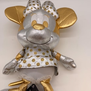 May include: A plush Minnie Mouse doll with a silver body, gold ears, and a gold nose. The doll wears a silver bow with gold polka dots and a dress with gold polka dots. The doll's arms and legs are silver and gold.