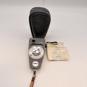 Puede incluir: Un fotómetro Minolta gris con una funda de cuero y un papel con texto japonés. El fotómetro tiene un dial circular con números y una correa de cuero. La funda es negra y tiene el logotipo de Minolta.