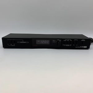May include: A black audio component with a digital clock display showing 12:00 AM. The device has various control knobs and buttons labeled "Input Tuning," "Output On," and "Pre Shock Control." The brand name is visible on the right side.