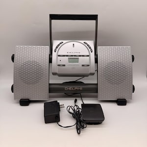 Può includere: Lettore CD Delphi argentato con due altoparlanti abbinati. Il lettore CD ha un display digitale e pulsanti di controllo. Gli altoparlanti sono rettangolari con una griglia metallica perforata. Include un adattatore di alimentazione nero e un telecomando nero.