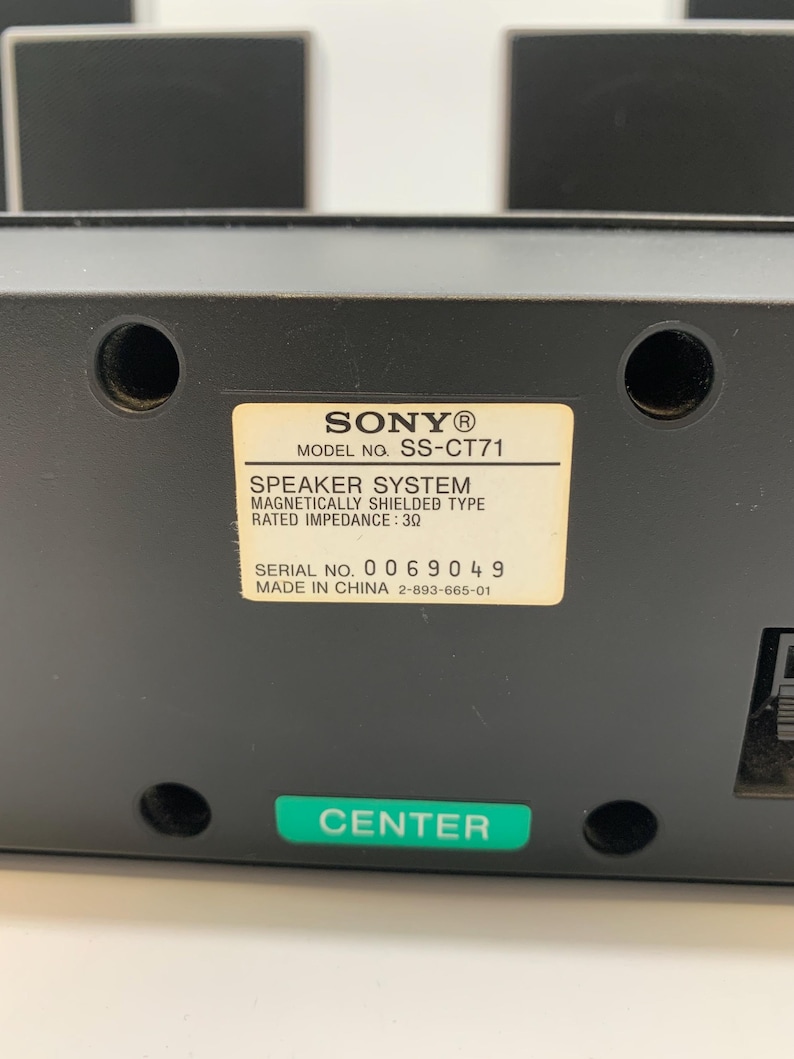 May include: A black Sony speaker system, model SS-CT71, featuring a center channel label. The back panel includes a white identification sticker with technical specifications, including a rated impedance of 3 ohms, and a green label marked CENTER.