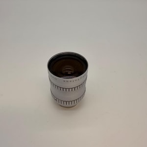May include: A silver Angenieux 10 mm camera lens. The lens has a black interior and a ridged exterior with the Angenieux brand name and other markings. The lens is cylindrical and sits on a white surface.