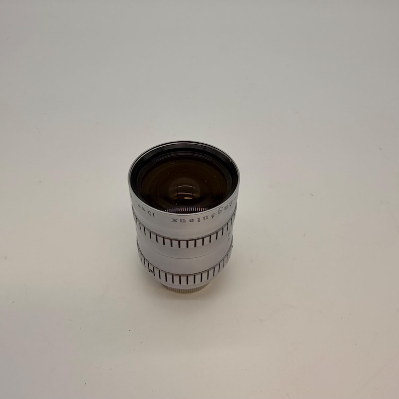 May include: A silver Angenieux 10 mm camera lens. The lens has a black interior and a ridged exterior with the Angenieux brand name and other markings. The lens is cylindrical and sits on a white surface.