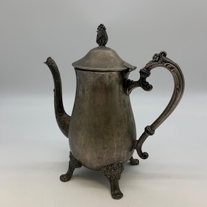 May include: An antique silver-colored teapot with a lid and ornate handle. The teapot has a curved spout and sits on three decorative feet. The handle and spout have detailed floral designs. The teapot is likely made of silver or a silver-plated metal.