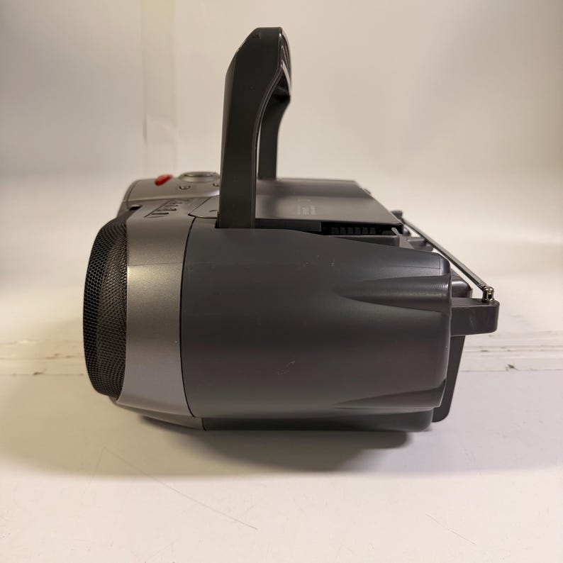 K&ouml;nnte beinhalten: Ein dunkelgrauer tragbarer Ghettoblaster mit einem markanten Tragegriff an der Oberseite, einem runden Lautsprechergitter an der Seite und einer ausziehbaren Metallantenne an der R&uuml;ckseite. Das Ger&auml;t verf&uuml;gt &uuml;ber ein Geh&auml;use aus Kunststoff.