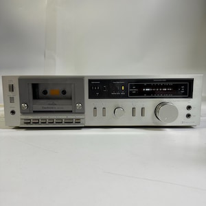May include: Silver Technics M225 cassette deck with a cassette tape inserted. The deck has various buttons, knobs, and a display screen. The Technics logo is visible on the front.