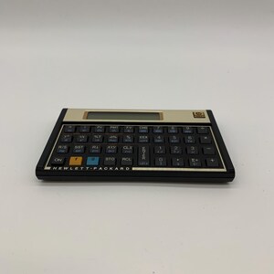 Könnte beinhalten: Ein Vintage-Taschenrechner von Hewlett-Packard in Schwarz und Beige. Der Rechner hat viele Tasten und ein Display. Der Markenname "HEWLETT-PACKARD" ist auf dem Rechner aufgedruckt.