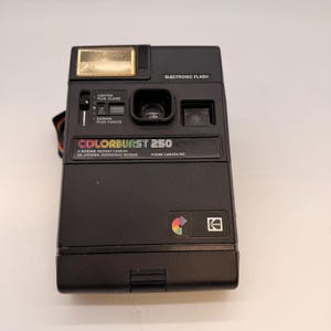 May include: Black Kodak Colorburst 250 instant camera with an electronic flash. The camera has a lens, viewfinder, and adjustment controls. The text "COLORBURST 250" and "KODAK" are visible on the front. A multi-coloured strap is attached.