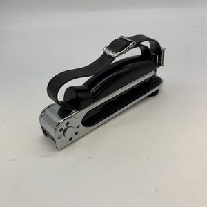 Peut inclure: Une pédale de vélo noire et chromée avec une sangle noire et une boucle argentée. La pédale a une forme rectangulaire avec un dessus incurvé et une finition chromée sur les côtés. La sangle est réglable.