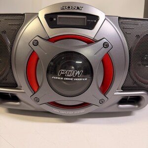 K&ouml;nnte beinhalten: Ein silbernes tragbares Sony-Audiosystem mit einem zentralen Power Drive Woofer mit rotem kreisf&ouml;rmigem Akzent. Das Ger&auml;t zeigt den Text PDW, Power Drive Woofer und Twin Amplifier System. Es enth&auml;lt einen digitalen Bildschirm und Lautsprechergitter.