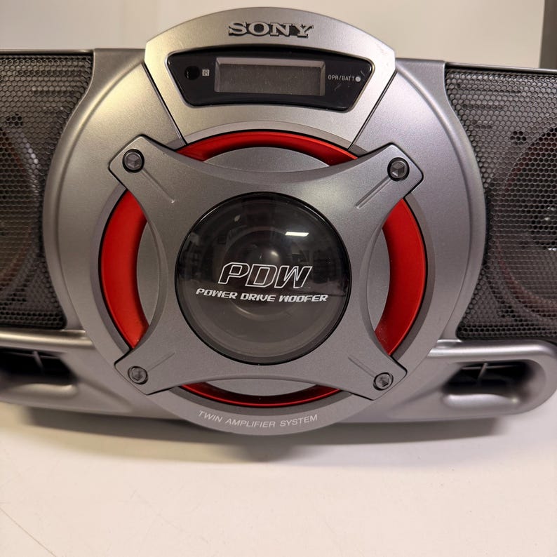 K&ouml;nnte beinhalten: Ein silbernes tragbares Sony-Audiosystem mit einem zentralen Power Drive Woofer mit rotem kreisf&ouml;rmigem Akzent. Das Ger&auml;t zeigt den Text PDW, Power Drive Woofer und Twin Amplifier System. Es enth&auml;lt einen digitalen Bildschirm und Lautsprechergitter.