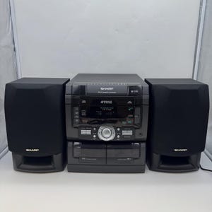 Puede incluir: Un equipo de música Sharp negro con dos altavoces a juego. El equipo de música tiene reproductor de CD, reproductor de casetes y radio. La pantalla muestra la hora. Los altavoces son rectangulares y tienen el logotipo de Sharp.