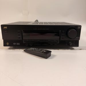 以下が含まれることがあります： 黒いJVCオーディオ/ビデオコントロールレシーバーとリモコン。レシーバーには大きなボリュームノブとディスプレイ画面があります。リモコンは黒色で、複数のボタンがあります。JVCのロゴが前面に表示されています。