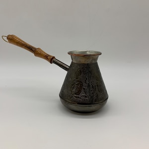 以下が含まれることがあります： 木製のハンドルが付いた、濃い色の金属製コーヒーポット。 ポットには、金属にエッチングされた装飾的な花の模様があります。 ハンドルは明るい色の木材でできており、金属製のブラケットでポットに取り付けられています。