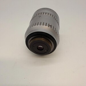 May include: A silver-colored camera lens with black accents. The lens has a cylindrical body with etched markings and a series of vertical lines. The lens is angled, revealing the dark interior of the lens.