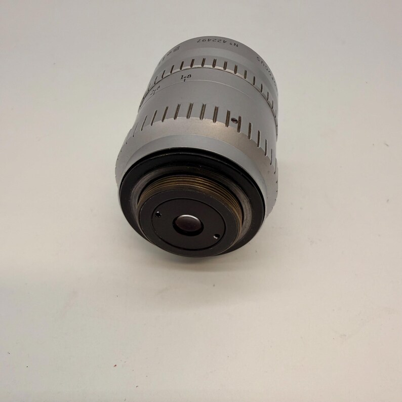 May include: A silver-colored camera lens with black accents. The lens has a cylindrical body with etched markings and a series of vertical lines. The lens is angled, revealing the dark interior of the lens.
