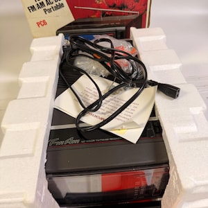 K&ouml;nnte beinhalten: Ein schwarzes und graues FM/AM 5" Farb-TV/FM-AM-Empfangsger&auml;t mit einem schwarzen Netzkabel und wei&szlig;er Verpackung. Auf der Box steht "FM/AM AC/DC Portable" und "PC6".