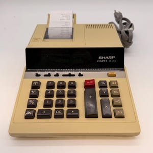 May include: A vintage Sharp Compet CS-2612 calculator with a beige body and black keys. The calculator has a red equals key and a paper roll outputting a printed receipt. The text "SHARP COMPET CS-2612" is visible.