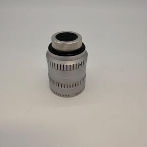 May include: A silver-colored camera lens with a black top and a ridged, cylindrical body. The lens has a series of vertical lines etched around its circumference and the text "Ang&eacute;nieux 10 mm" is visible. The lens is designed for professional photography.