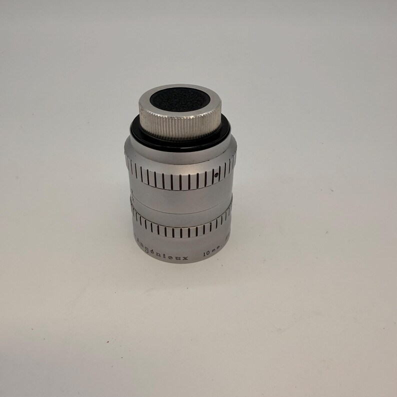 May include: A silver-colored camera lens with a black top and a ridged, cylindrical body. The lens has a series of vertical lines etched around its circumference and the text "Ang&eacute;nieux 10 mm" is visible. The lens is designed for professional photography.