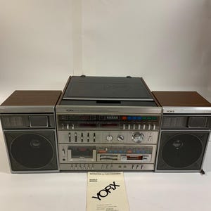 Puede incluir: Sistema estéreo Yorx vintage con altavoces, reproductor de casetes y radio. El sistema tiene una cara plateada con detalles de vetas de madera marrón. Un manual con el logotipo de Yorx está al frente.