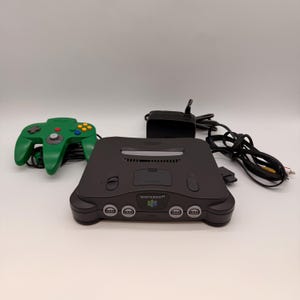 May include: A black Nintendo 64 game console with a green controller and power adapter. The controller has a directional pad, buttons, and a joystick. The console has the Nintendo 64 logo.