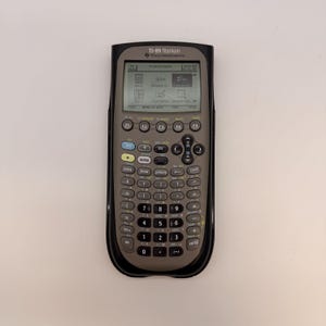 May include: A TI-89 Titanium graphing calculator with a black protective case. The calculator has a gray keypad with numerous buttons, including function keys, number keys, and navigation buttons. The screen displays text and graphics. The Texas Instruments logo is visible.