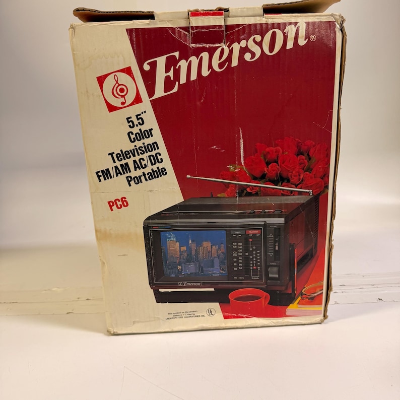 K&ouml;nnte beinhalten: Ein Emerson 5,5" Farbfernseher in der Originalverpackung. Die Box ist rot und wei&szlig; mit dem Emerson-Logo und Produktinformationen. Der tragbare Fernseher hat ein schwarzes Geh&auml;use und einen kleinen Bildschirm, der eine Stadtlandschaft zeigt.