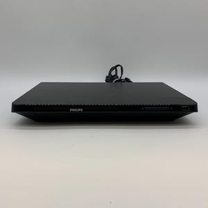 Può includere: Un lettore DVD Philips nero con un design elegante e rettangolare. La parte anteriore del dispositivo presenta il marchio "PHILIPS" in lettere bianche. Un cavo di alimentazione è collegato sul retro.