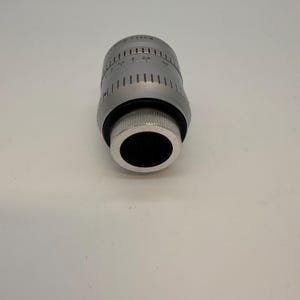May include: A silver-colored camera lens with black accents. The lens has measurement markings and a ridged grip. The lens is cylindrical with a black opening at the end.