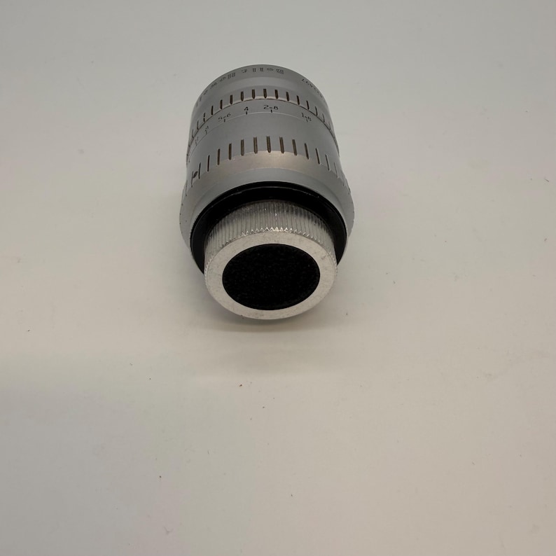 May include: A silver-colored camera lens with black accents. The lens has measurement markings and a ridged grip. The lens is cylindrical with a black opening at the end.