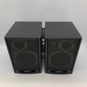 May include: Two black Sony satellite speakers positioned side-by-side. Each rectangular speaker features a front-facing mesh grille covering two circular drivers, with the word SAT printed near the top and the Sony logo at the bottom.