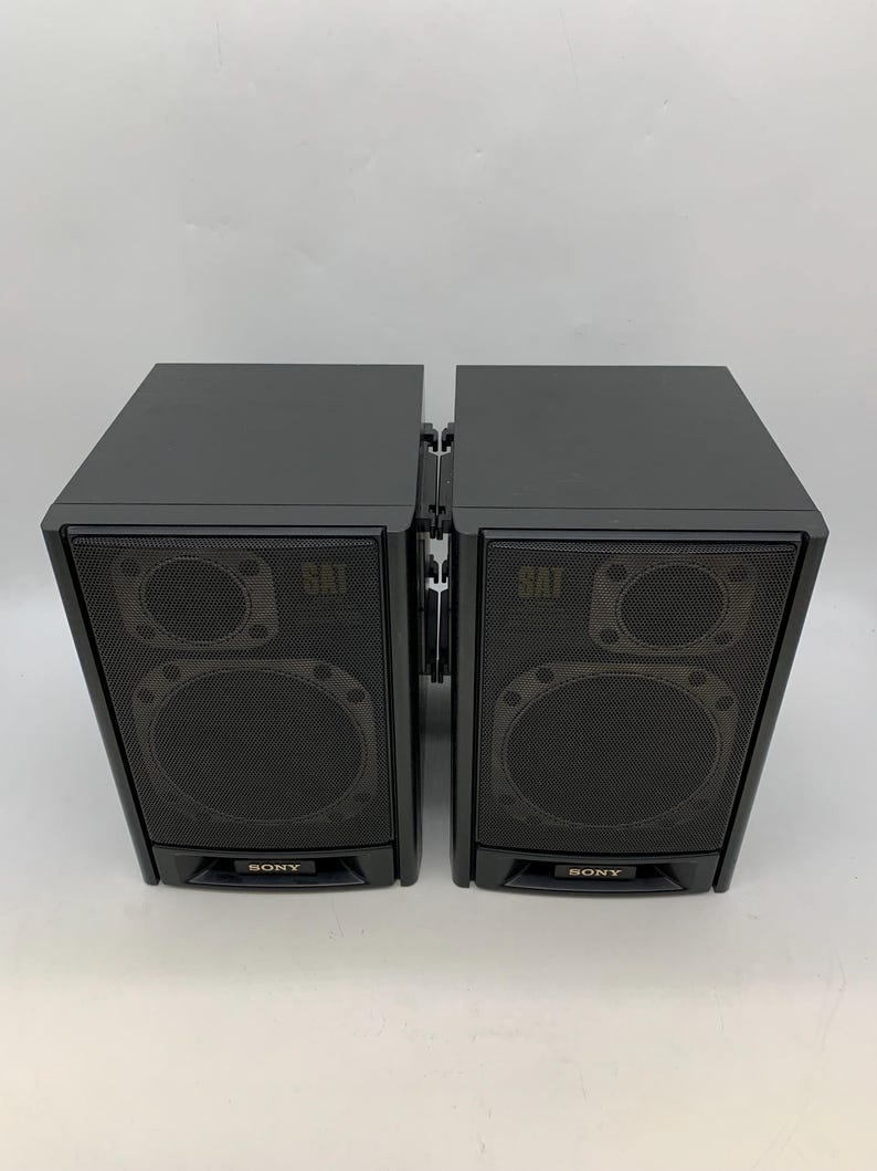 May include: Two black Sony satellite speakers positioned side-by-side. Each rectangular speaker features a front-facing mesh grille covering two circular drivers, with the word SAT printed near the top and the Sony logo at the bottom.