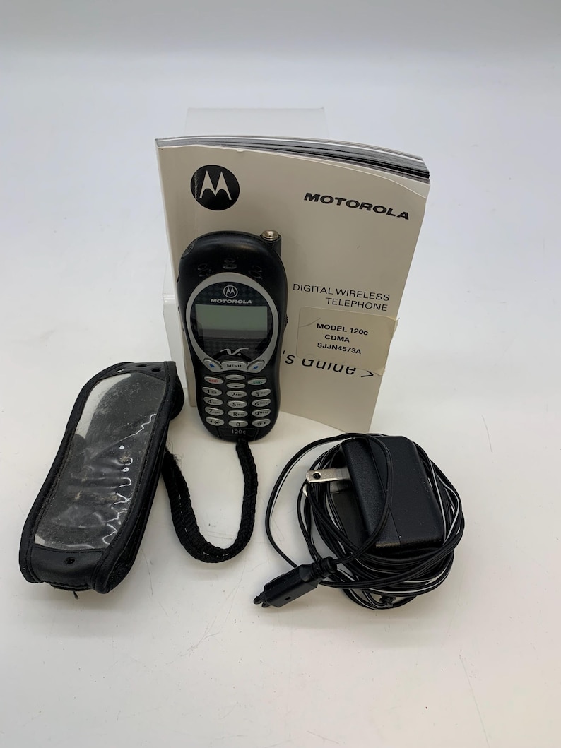 May include: A black Motorola 120c digital wireless telephone with a screen and keypad. Included are a black carrying case, charger, and a user guide. The Motorola logo is visible on the phone and the guide.