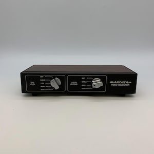 Op de afbeelding: Een zwarte Archer video selector met twee draaiknoppen en witte tekst. De knoppen zijn gelabeld met "TV Viewing" en "VTR Recording". Het apparaat heeft ingangen voor antenne, kabel en hulpapparatuur.