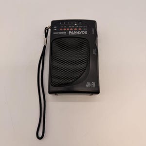 May include: A black Panavox AM/FM pocket radio with a black wrist strap. The radio features a speaker grill, tuning dial, and the words "AM FM" printed on the front. The radio is rectangular and compact.
