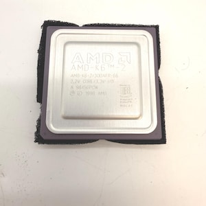 Puede incluir: Un procesador AMD-K6 TM-2 plateado con espuma protectora negra. El procesador tiene el logotipo de AMD y el texto "AMD-K6 TM-2", "AMD-K6-2/300AFR-66" y "1998 AMD".