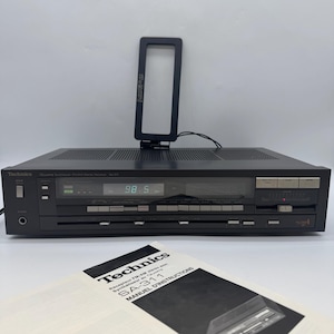 Könnte beinhalten: Schwarzer Technics SA-311 Stereo-Receiver mit digitaler Anzeige von 98,5. Er hat eine schwarze Antenne und ein Handbuch im Vordergrund. Das Handbuch trägt die Aufschrift "Technics SA-311 MANUAL D'INSTRUCTIONS".