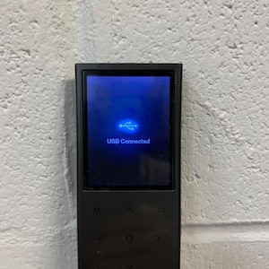 Puede incluir: Un reproductor multimedia digital negro con una pantalla que muestra "USB Connected" y un símbolo USB. El dispositivo tiene botones de control y está conectado a un cable negro. El fondo es una pared blanca texturizada.