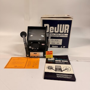 May include: A black DeJUR Specto 500 Dual 8mm Movie Editor with a viewing screen, two reel arms, and a box. The box is white and navy blue with the text "DeJUR" and "Spectator". Also included are instruction sheets and a brochure.