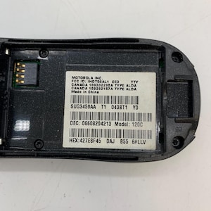 May include: A close-up of the inside of a black Motorola 120C phone. The phone's internal components are visible, including a circuit board and a label with technical specifications. The label includes the FCC ID, model number, and other regulatory information.