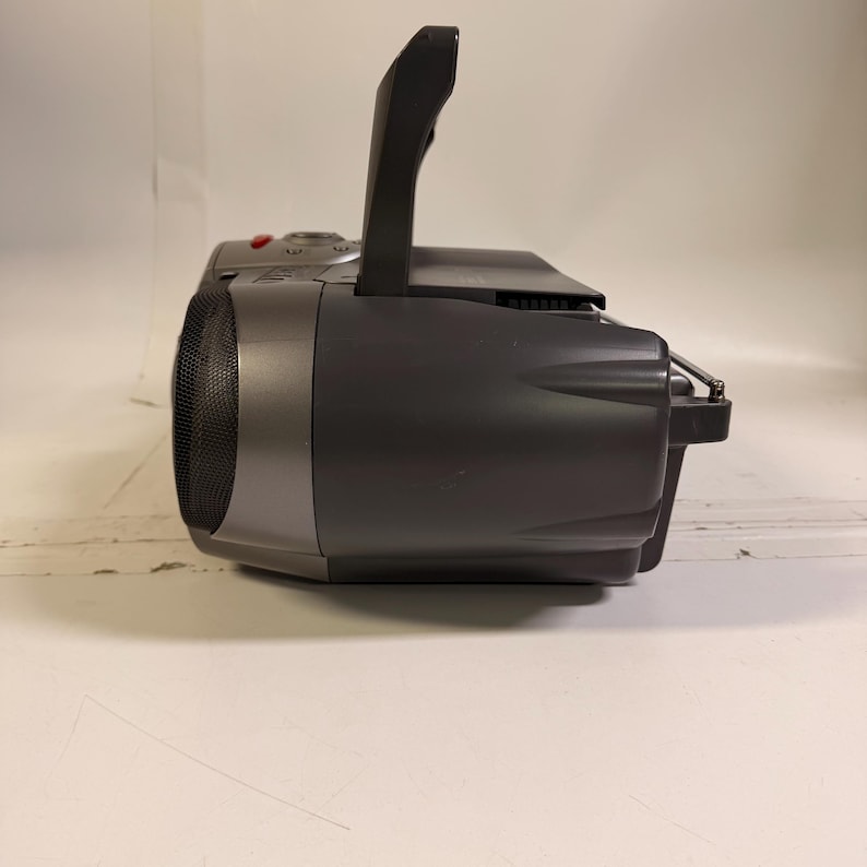 K&ouml;nnte beinhalten: Ein dunkelgraues tragbares Radio mit CD-Player, ausgestattet mit einem seitlichen Lautsprecher mit schwarzem Netzgitter, einem aufrechten Tragegriff an der Oberseite und einer d&uuml;nnen Metallantenne, die aus der R&uuml;ckseite des Geh&auml;uses ragt.