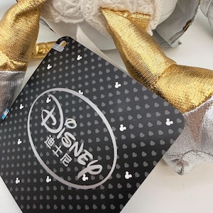 May include: A close-up of a Disney-branded plush toy with gold and silver accents. The tag features the Disney logo and Chinese characters on a black background with white Mickey Mouse icons.