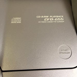 K&ouml;nnte beinhalten: Ein grauer CD-Radio-Kassettenrekorder mit dem Text CD-R/RW PLAYBACK CFD-G55 CD RADIO CASSETTE-CORDER auf der Oberseite. Das Ger&auml;t verf&uuml;gt &uuml;ber ein Compact Disc Digital Audio-Logo und eine runde Taste mit der Aufschrift PUSH OPEN/CLOSE.
