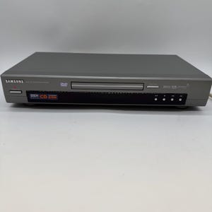 May include: A grey Samsung DVD player with a disc tray and a black control panel. The player has the DVD logo and the words "MP3 CD-R/RW" on the front. The device is on a white surface.