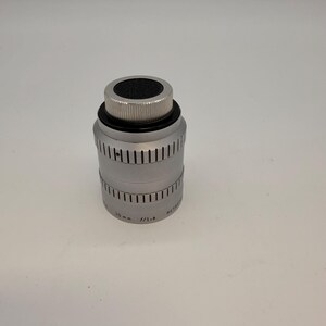 May include: A silver-colored camera lens with a black top and ridged metal detailing. The lens has a cylindrical body with vertical grooves and text that reads "10 mm f/1.6".