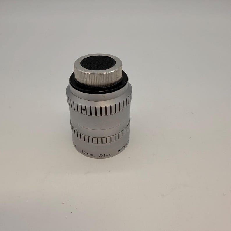 May include: A silver-colored camera lens with a black top and ridged metal detailing. The lens has a cylindrical body with vertical grooves and text that reads "10 mm f/1.6".