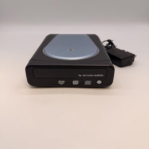 Può includere: Masterizzatore DVD HP dvd writer dvd630e nero e grigio con parte superiore blu chiaro. La parte anteriore del dispositivo presenta i simboli DVD, RW e disco. Il testo "hp dvd writer dvd630e" è stampato sulla parte anteriore. Include un adattatore di alimentazione nero.