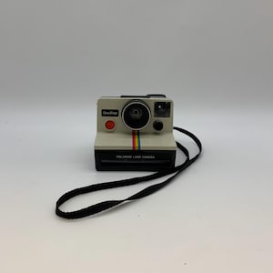 Peut inclure: Un appareil photo Polaroid OneStep Land vintage, de couleur crème et noir, avec une bande arc-en-ciel. L'appareil photo a un bouton rouge et une sangle noire. Les mots "OneStep" et "Polaroid Land Camera" sont imprimés.
